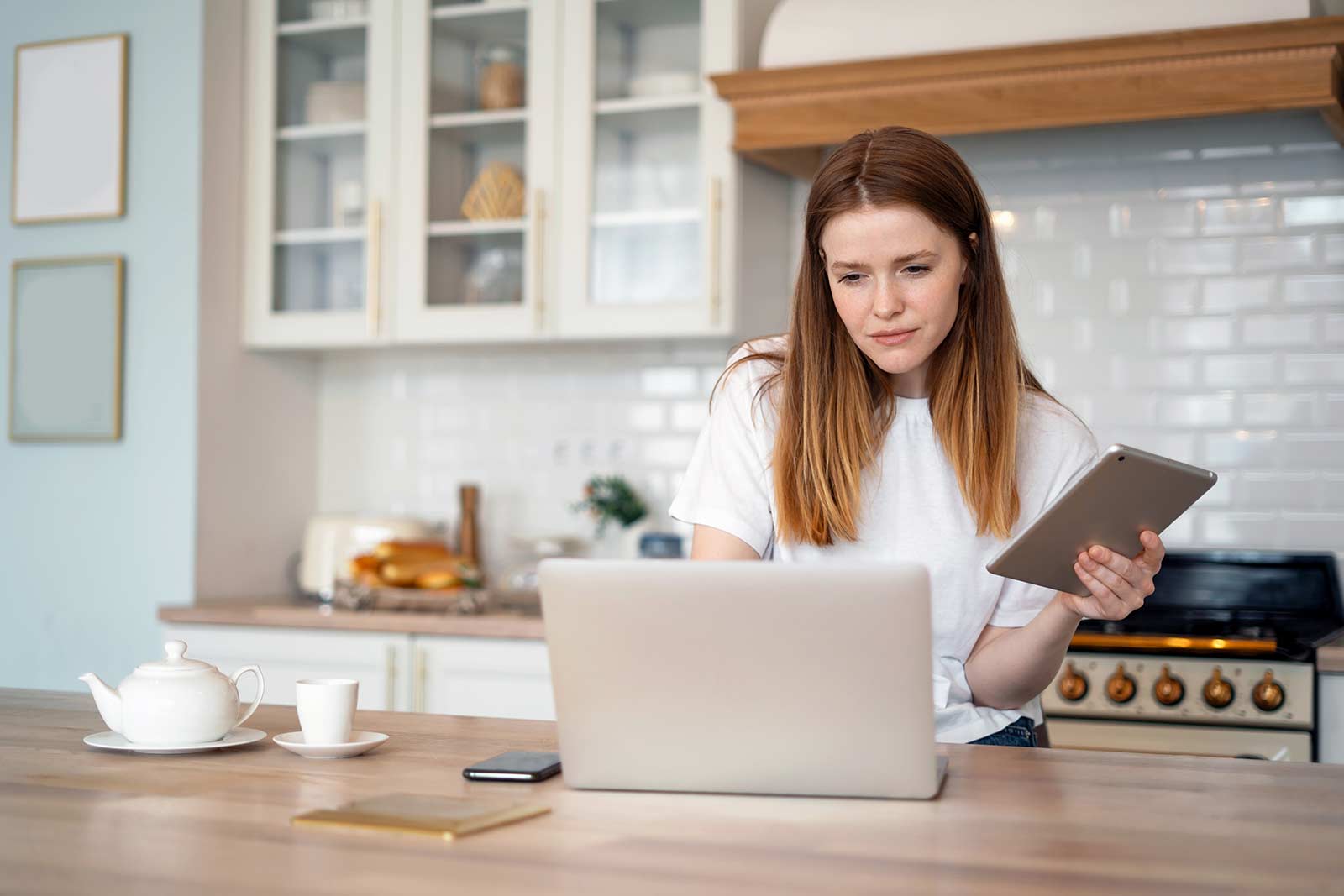 Young woman in her kitchen using her laptop and tablet to do online research